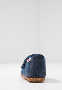 Chaussons en laine bleue avec une sangle en velcro, un talon en suède bleu foncé contrastant et une semelle en caoutchouc. Présente une petite étiquette sur le côté.