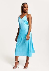 A blue satin spaghetti strap dress with a V-neck, flowing midi length, and a slight high-low hem. Worn with silver heeled sandals.