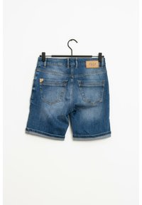 Pantalones cortos de mezclilla azul con dobladillos enrollados colgando de una percha negra, mostrando los bolsillos traseros y una etiqueta de cuero "PULZ JEANS" en la cintura.