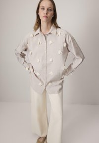 Camisa de botones gris claro con bordado floral blanco en el frente, combinada con pantalones anchos color crema, sobre un fondo neutro.