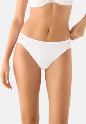 Mujer con ropa interior estilo bikini blanca con paneles laterales de malla y un sujetador blanco a juego, de pie contra un fondo claro y sencillo.