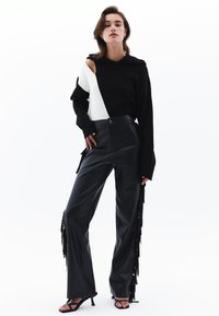 Schwarzer und weißer asymmetrischer Pullover mit Kapuze, kombiniert mit hoch sitzenden schwarzen Lederhosen mit Fransenverzierungen und Absatzsandalen.