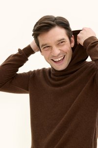 Brauner Strickpullover mit hohem, umgeschlagenem Kragen; weiche Textur; eng zulaufende Ärmel; getragen von einem Mann, der die Kapuze über seinen Kopf zieht.