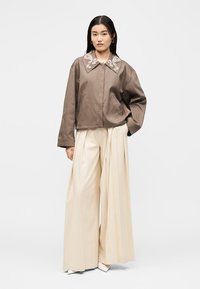 Γυναίκα που φοράει ένα φαρδύ καφέ σακάκι με κεντητό γιακά, κρεμ παντελόνια σε φαρδιά πλισαριστή γραμμή και λευκά μυτερά παπούτσια, στέκεται μπροστά σε απλό φόντο.