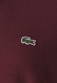 Burgundy stickad tyg med en grön krokodil-logotyp med röda accenter, omringad av vitt. Texturen verkar slät och mjuk.