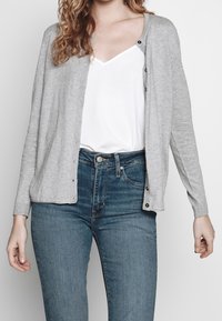 Grå cardigan i mjukt stickat tyg, med rund halsringning, knappstängning och långa ärmar, i kombination med en vit topp och blå jeans.