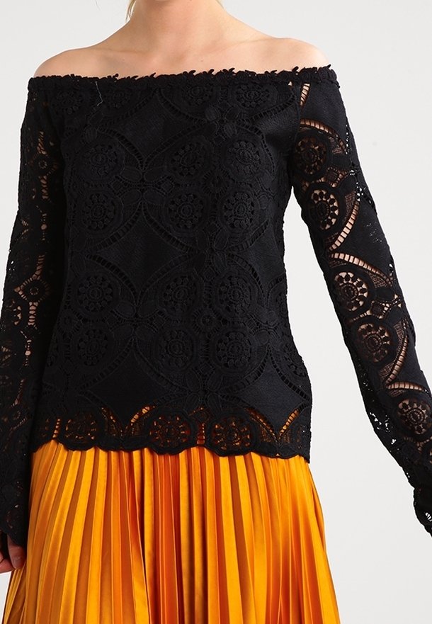 Haut en dentelle noir, décolleté asymétrique avec des manches longues ornées de motifs floraux complexes, associé à une jupe plissée orange.