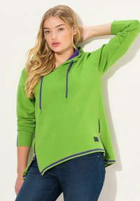 Felpa verde con cappuccio e dettagli viola, orlo asimmetrico, tasche con zip e polsini a coste. Materiale morbido con una vestibilità rilassata.