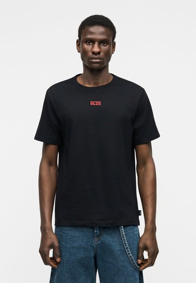 Άνδρας που φοράει μαύρο κοντομάνικο μπλουζάκι με κόκκινο λογότυπο "GCDS" στο στήθος και μπλε τζιν παντελόνι με ορατή αλυσίδα στη θηλιά της ζώνης.