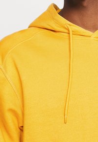 Persona que lleva un sudadera con capucha de color amarillo brillante, con cordones visibles y tejido texturizado, en vista cercana.