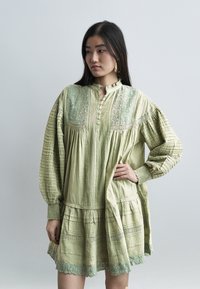 Νεαρή γυναίκα με μακριά σκουρόχρωμα μαλλιά, φορώντας ένα φαρδύ, ανοιχτό πράσινο φόρεμα με κεντημένες λεπτομέρειες και κουμπιά, στέκεται με το ένα χέρι στη μέση.