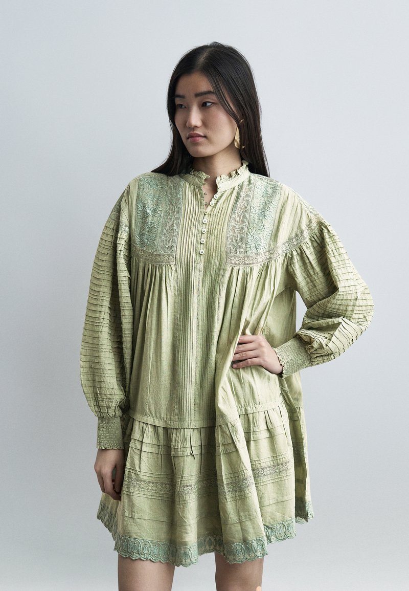 Νεαρή γυναίκα με μακριά σκουρόχρωμα μαλλιά, φορώντας ένα φαρδύ, ανοιχτό πράσινο φόρεμα με κεντημένες λεπτομέρειες και κουμπιά, στέκεται με το ένα χέρι στη μέση.