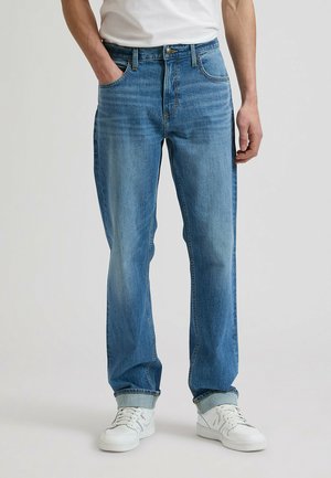 Man draagt lichtblauwe rechtvallende spijkerbroek met omgeslagen zoom, witte sneakers en een wit t-shirt, staat met één hand in de zak.