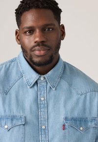 Hele sinine teksasärk, millel on klassikaline nööbitav eesmine osa, terav kaelus ja kaks rinnataskut eristuva õmblusega.