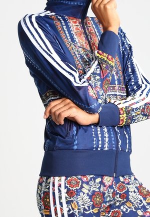 Veste zippée bleu marine avec des motifs floraux et des rayures blanches sur les manches. Manches côtelées et ourlet, avec une poche latérale zippée.