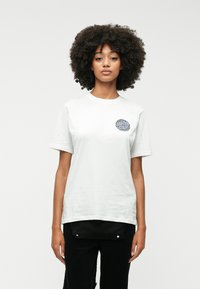 Biela bavlnená tričko s krátkymi rukávmi, okrúhlym výstrihom, so zaobleným modrým grafickým motívom na ľavej hrudi. Dole sú oblečené čierne nohavice.