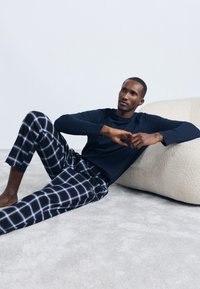 Mornarska majica s dugim rukavima u kombinaciji s udobnim hlačama na vezanje u crno-bijelom kariranom uzorku. Mekana tekstura i opušteni kroj. Svijetlo siva pozadina.