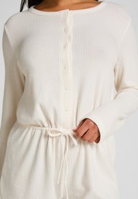 Prenda de manga larga blanca de canalé con botones en la parte delantera, que presenta una cintura ajustable con cordón y una textura suave.