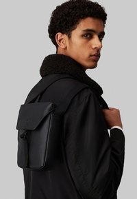 Mochila negra texturizada con cierre de solapa, llevada al hombro. Presenta una correa ajustable y un logo sutil en la parte frontal. Acabado suave y mate.