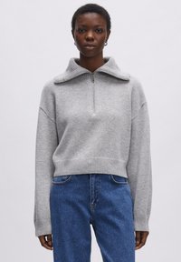 Grijze gebreide trui met een hoge kraag, half-rits aan de voorkant en geribbelde zoom en manchetten. Gepaard met blauwe jeans.