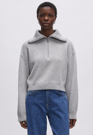 Grijze gebreide trui met een hoge kraag, half-rits aan de voorkant en geribbelde zoom en manchetten. Gepaard met blauwe jeans.