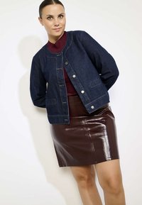 Veste en denim bleu foncé avec un col rond, fermeture à boutons et deux poches à l'avant, portée sur un haut côtelé bordeaux et une jupe en cuir brun brillant.