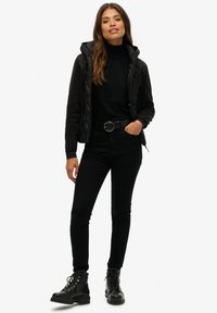 Veste matelassée noire avec capuche, portée sur un col roulé noir. Un pantalon skinny noir et des bottes de combat à lacets complètent la tenue.