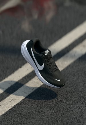 Fekete-fehér Nike futócipő lebeg az aszfalt fölött, alatta fehér útjelzésekkel.