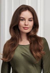 Junge Frau mit langen, gewellten braunen Haaren, die eine grüne Satinbluse und kleine goldene Creolen trägt, vor einem weißen, vertäfelten Wandhintergrund.