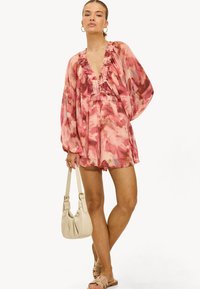 Vrouw in een roze gemêleerde romper met lange puffmouwen, met een beige handtas in de hand en beige sandalen, tegen een witte achtergrond.