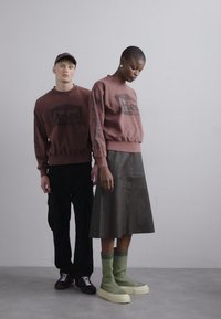Brun sweatshirt med logo, parret med en mørkegrøn læderskirt. Modellen har grønne strikkede sokker og kraftige cremede sko på; en anden model i sorte bukser.