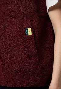 Vinröd fleeceväst med en mjuk, strukturerad yta, utrustad med en framficka och en färgglad etikett med "PCS" och en hjärtdesign.