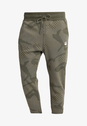 Pantalon de jogging vert olive avec taille à cordon, présentant un motif camouflage pixelisé et à carreaux ainsi qu'un petit logo blanc sur la cuisse droite.