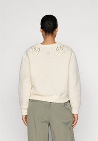Sudadera color crema en un material suave y texturizado, con un yugo bordado de patrón de diamante y un parche de logo cuadrado en la parte posterior del cuello.