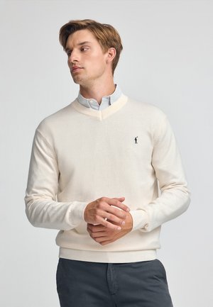 Suéter de color crema con cuello en V, hecho de una tela suave, que presenta un pequeño logo bordado en el lado izquierdo del pecho y puños y dobladillo acanalados.