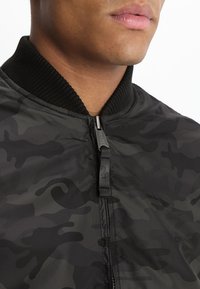 Gros plan sur une personne portant une veste zippée camouflage noire et gris foncé avec un col côtelé.