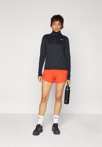 Haut de sport zippé noir, shorts orange, baskets noires. Tient une bouteille d'eau noire, chaussettes blanches avec logo, et un fond neutre.