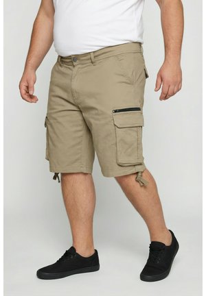 Hombre con pantalones cortos cargo beige con bolsillos laterales y zapatillas negras, de pie contra un fondo claro.