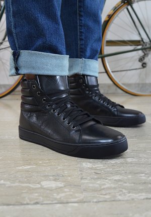 Zapatillas altas negras de cuero texturizado, con diseño de cordones, puntera redonda, suela de goma y soporte acolchado para el tobillo. Se pueden ver jeans de mezclilla.