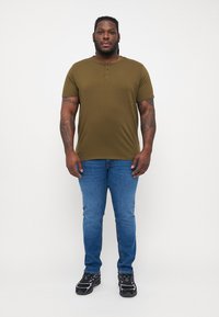 Olívazöld, rövid ujjú henley ing három gombbal, kék, slim-fit farmerral és fekete sportcipővel párosítva.