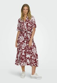 Abito floreale bordeaux con motivi a fiori bianchi, maniche corte e vita annodata. Realizzato in tessuto leggero, arriva a metà polpaccio, abbinato a sneaker bianche.