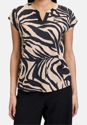 Vrouw draagt een beige top met korte mouwen met zwarte abstracte zebraprinten en een ingezakte halslijn, gecombineerd met een zwarte broek.