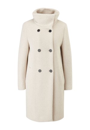 Personne portant un manteau croisé beige clair, un haut blanc, un pantalon noir taille haute avec des fentes latérales et des baskets blanches, debout.