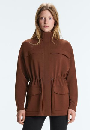 Nő barna, magas nyakú kabátot visel, amelynek nagy első zsebei és húzózsinóros derékvonala van, egyszínű világos háttér előtt áll.