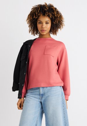 Göndör hajú nő rózsaszín hosszú ujjú zsebes felsőt, világoskék farmert és vállára dobott fekete dzsekit visel.