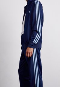Person i mörkblå Adidas-träningsoverall med ljusblå tre-stripes detaljer på ärmarna och byxorna, stående mot en enkel vit bakgrund.