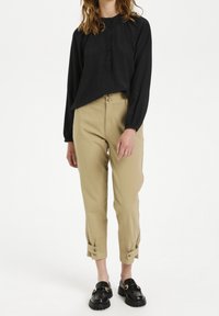 Svart långärmad tröja med henley-krage och knappar; beiga tapered byxor med uppvikta cuffs; svarta loafers med gulddetaljer.