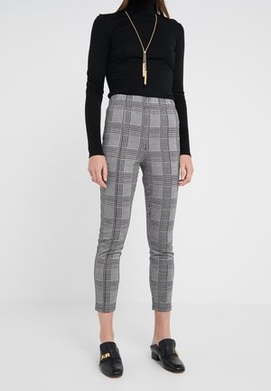 Femme portant un haut à manches longues noir, un collier en or, un pantalon gris à carreaux coupe slim et des chaussures slip-on noires avec des accents dorés, debout sur un fond blanc.