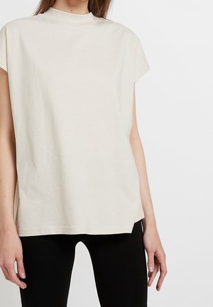 Beige short-sleeve top with a high neckline, made of soft cotton, featuring a relaxed fit and straight hem. Worn with black leggings.