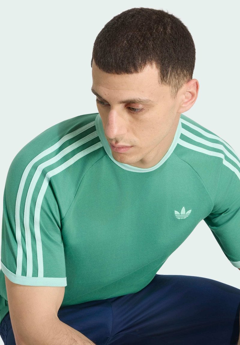 Homme portant un t-shirt vert Adidas avec trois bandes blanches aux épaules, regardant vers le bas, assis devant un fond clair.